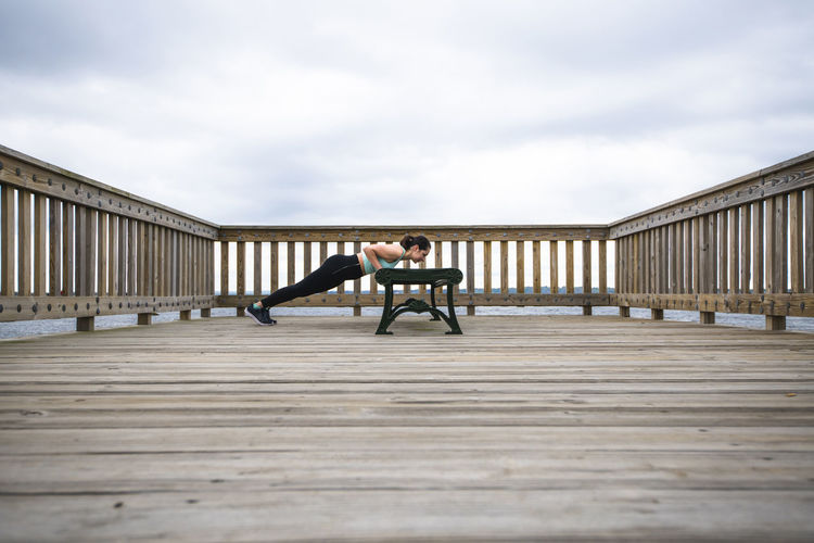 Side view of woman doing push-ups on bench at pier against cloudy sky