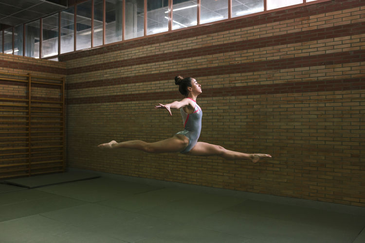 Full length of female gymnast with legs apart and arms outstretched jumping while exercising at gym