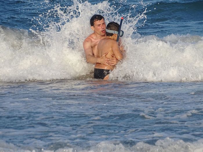Shirtless father and son standing amidst wave in sea