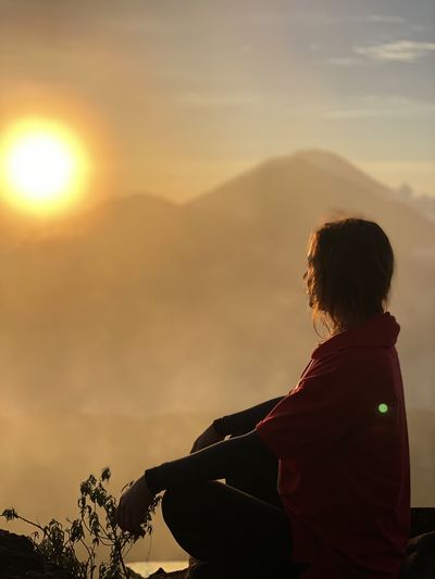 Side view of man siting on mountain against sky during sunset