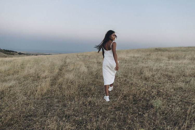 Woman wearing white dress walking in meadow in front of sky