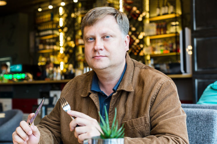 An adult blond man is having lunch in a cafe in a brown jacket.