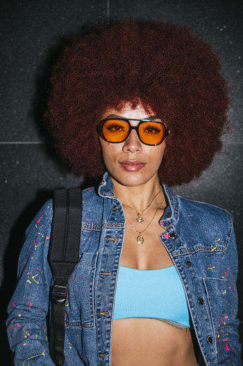 Confident female with afro hairstyle and trendy outfit and sunglasses looking at camera while standing on black background with backpack in evening time