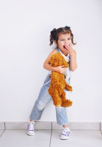 Smiling girl with teddy bear standing against wall at home