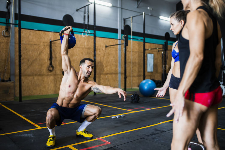 Male fitness instructor lifting kettlebell and practicing squats while assisting women in gym