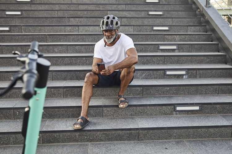 Smiling mature man with e-scooter sitting on stairs using cell phone