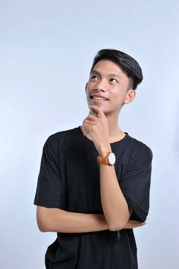 Handsome young asian boy wearing black t-shirt and wristwatch with hand on chin thinking about question, pensive expression. smiling with thoughtful face. doubt concept