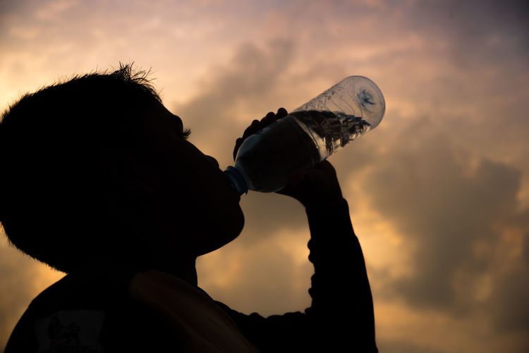 Close-up portrait of silhouette boy drinking water against sky during sunset