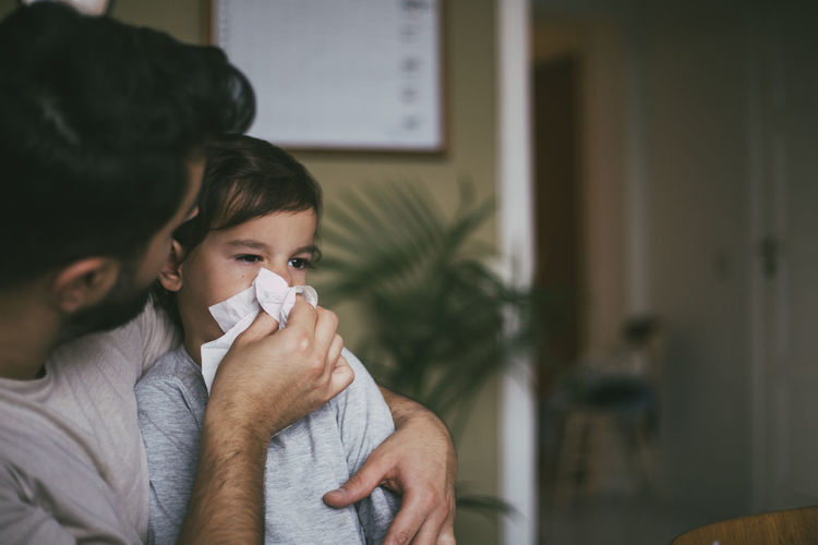 Father blowing nose of sick son at home
