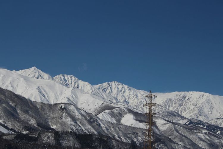 Low angle view of electricity pylon by snow covered mountains against clear blue sky