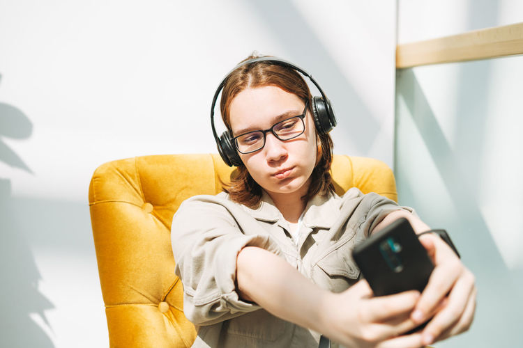 Young teenager girl student in glasses using mobile, listen music in headphones in yellow chair 