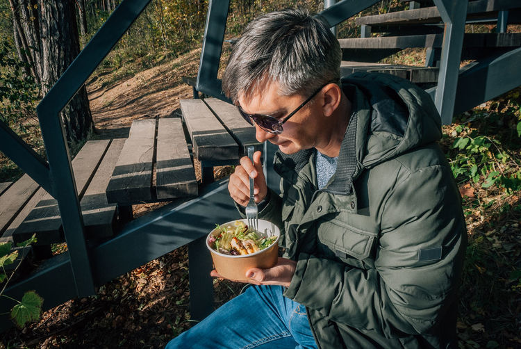 Man dines on a salad from disposable paper bowl in the open air during a walk in nature in the fall