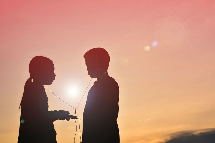 Silhouette friends listening music through headphones against sky during sunset
