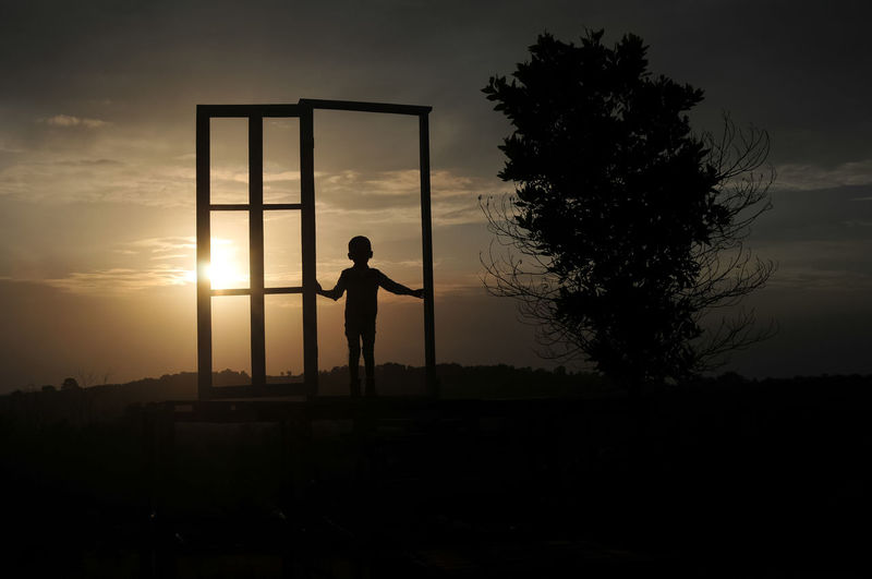 Silhouette boy walking at gate on field against sky during sunset