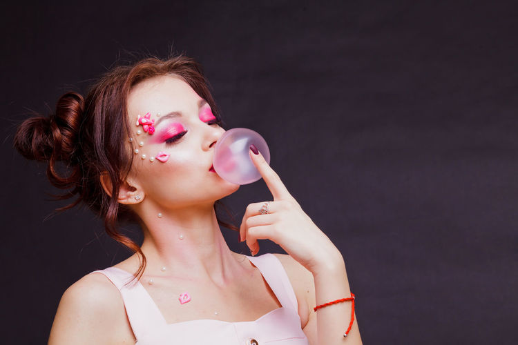 Beautiful young woman with make-up blowing bubble gum against gray background