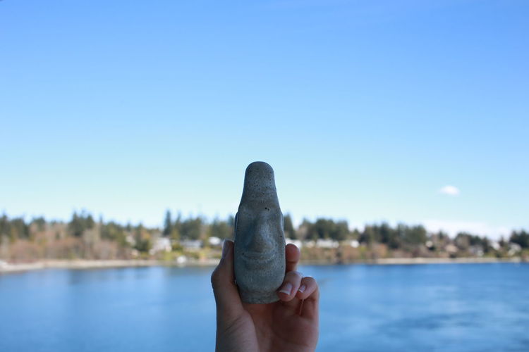 Cropped hand of woman holding face shape stone against lake ad sky