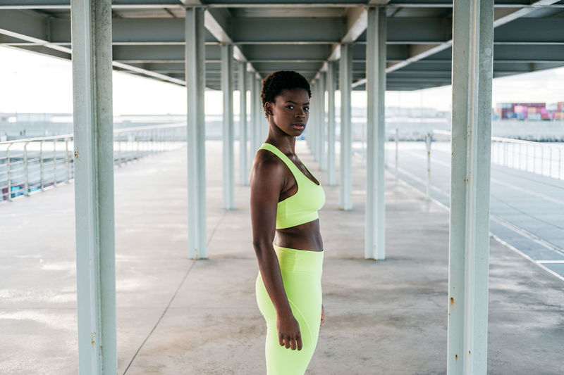 From below african american adult sportswoman in vibrant green activewear focusing standing alone along waterfront among metal columns under roof