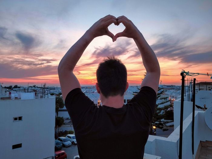 Rear view of man standing on rooftop against sunset sky making a love sign with his hands