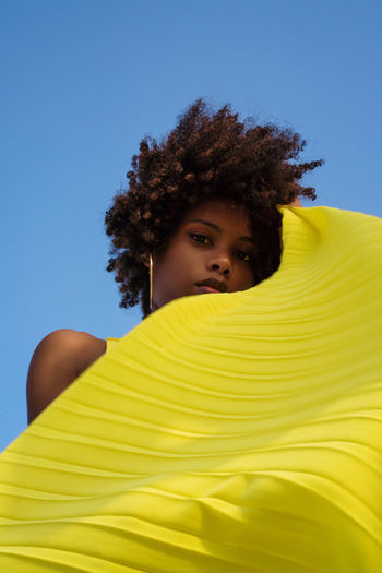 Low angle shot beautiful black woman with afro wearing yellow dress against blue sky