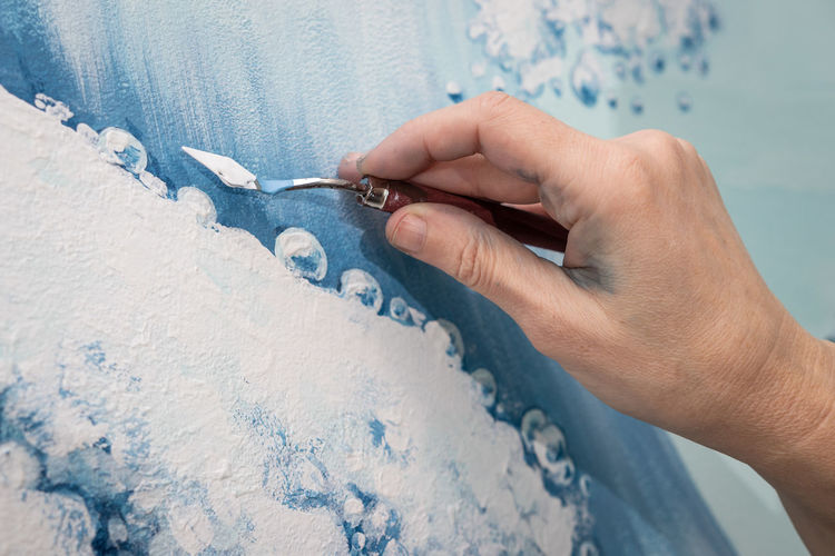 The hand of a woman artist draws an image of a sea wave using a palette knife, close uo