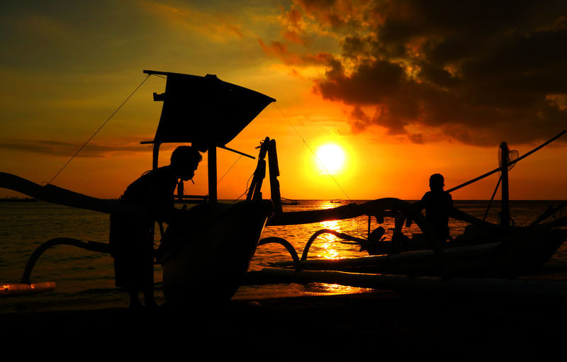 Silhouette boats and men at beach against orange sky during sunset