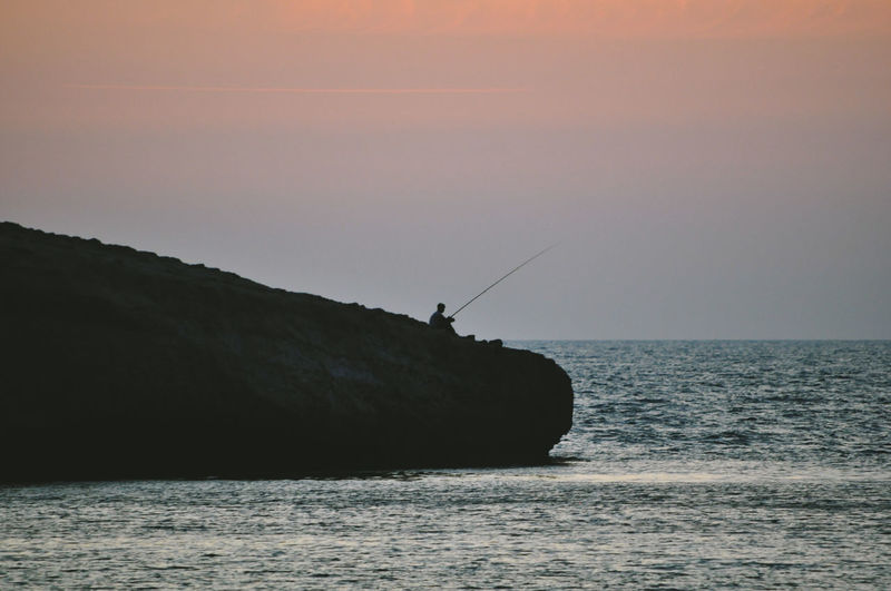 Silhouette fishing rod on rock by sea against sky