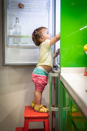 Side view of cute little child standing on stool and taking food from open refrigerator in cozy kitchen at home