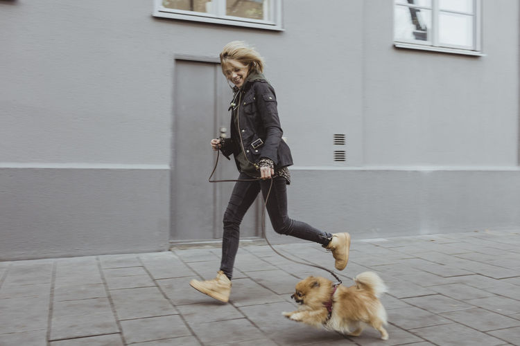 Full length side view of happy woman running with her pomeranian dog on footpath by building in city