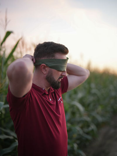 A bearded young man stands in a cornfield and covers his eyes with a corn leaf.