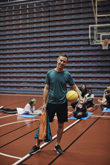 Full length portrait of smiling male coach standing with basketball against students at sports court