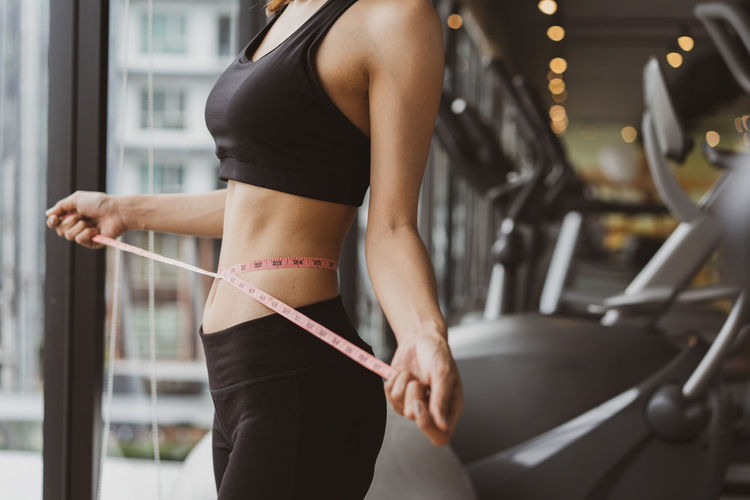 Midsection of woman measuring waist while standing in gym