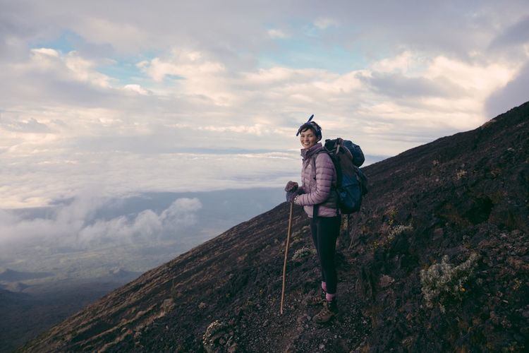 Portrait of smiling hiker standing on mountain against cloudy sky