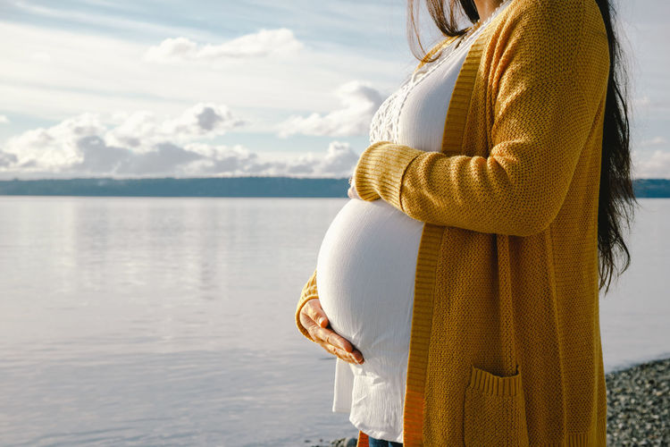 Midsection of pregnant woman standing on shore at beach against cloudy sky during sunset