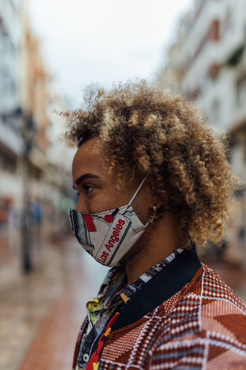 Side view of modern stylish african american male with curly hair dressed in trendy colorful informal clothes and protective mask standing against blurred urban environment and looking away