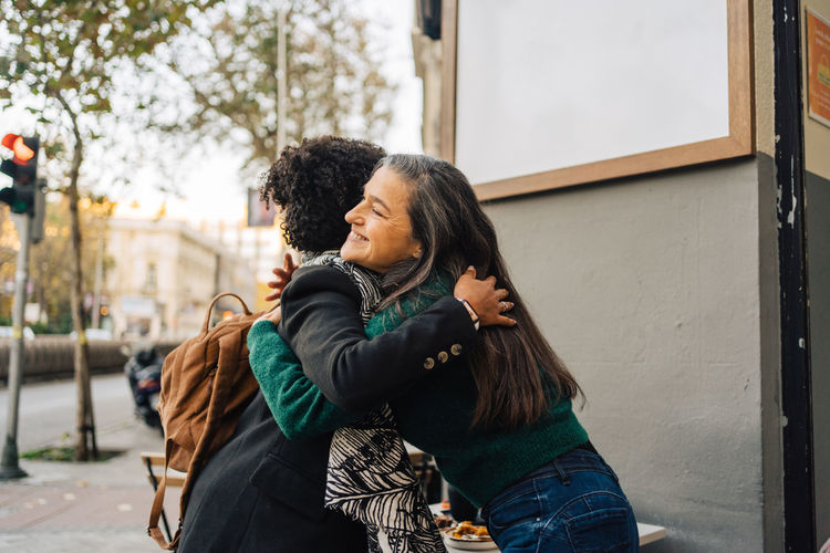 Side view of smiling mature hispanic woman embracing black curly haired girlfriend while greeting each other on street