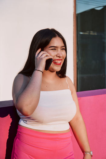 Happy overweight ethnic woman with long black hair and red lips smiling and looking away while leaning on border near pink wall and answering phone on call on city street