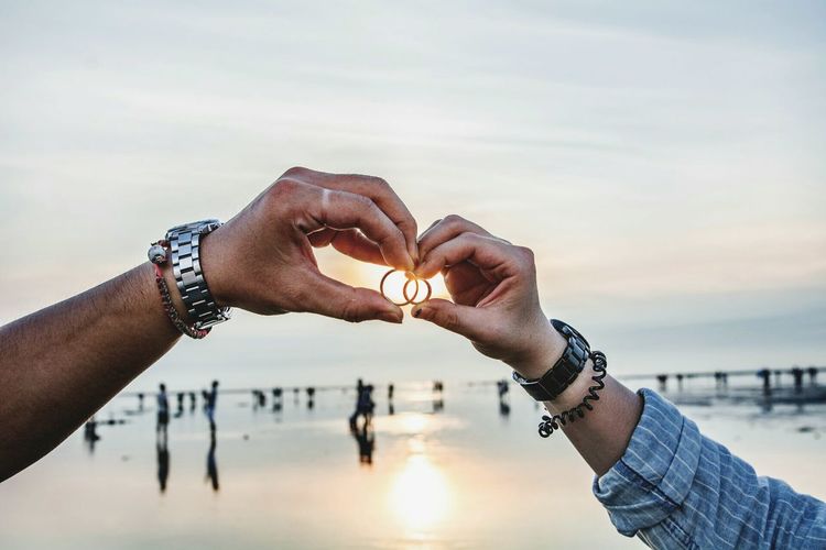 Cropped image of couple holding rings at beach against sky