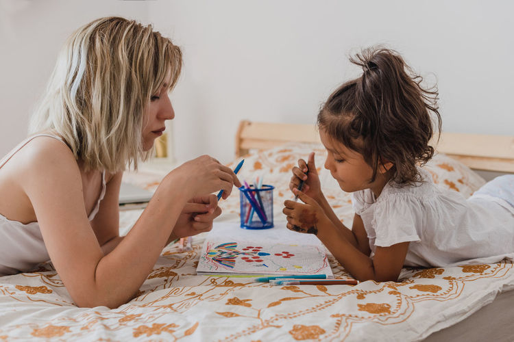 Caucasian mom and hispanic daughter painting while they are lying in the bed.