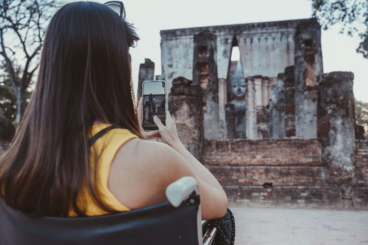 Rear view of woman photographing old ruins while sitting on wheelchair
