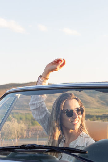 Vertical portrait of young beautiful woman with arm raised having fun on a convertible car 