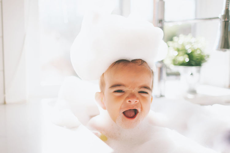 Cute happy baby boy taking bubble bath in kitchen sink at home
