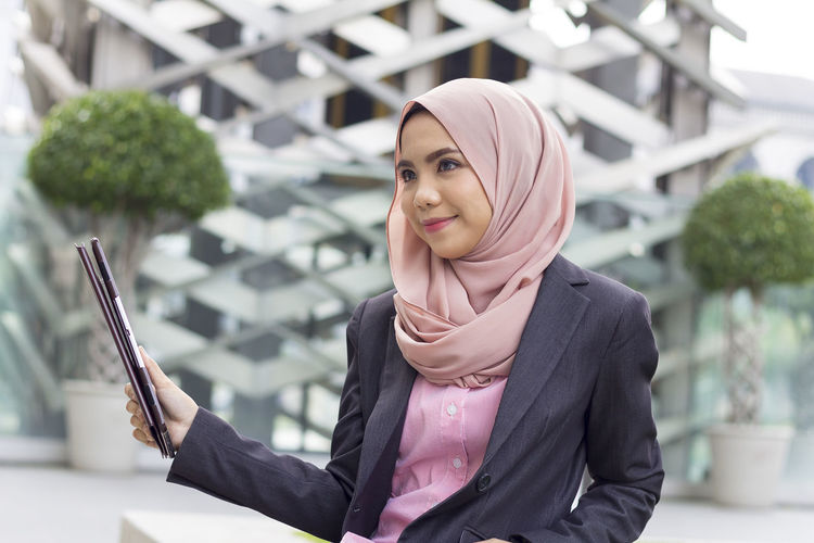 Thoughtful businesswoman holding digital tablet while standing against modern office building