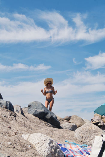 Full length of shirtless woman with bikini on rock at beach against sky