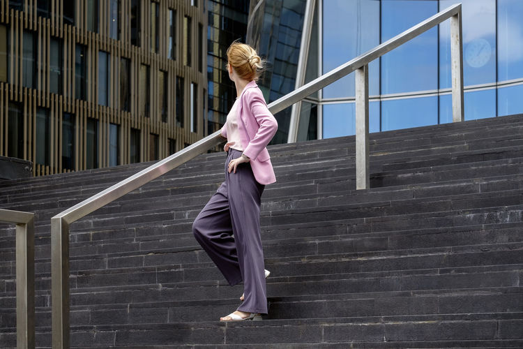 Young blonde with  stands on steps near modern office building in full growth, half-turned to camera