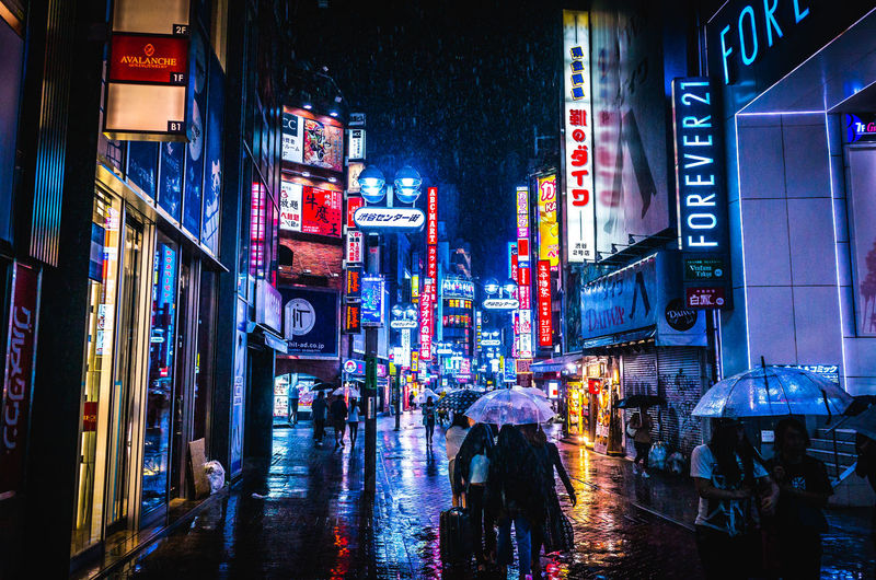 People on street amidst illuminated buildings during rainy season at shibuya