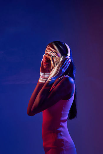Charming brazilian female listening to music in wireless headphones while standing with eyes closed in obscure studio on dark background with shadow of light on face
