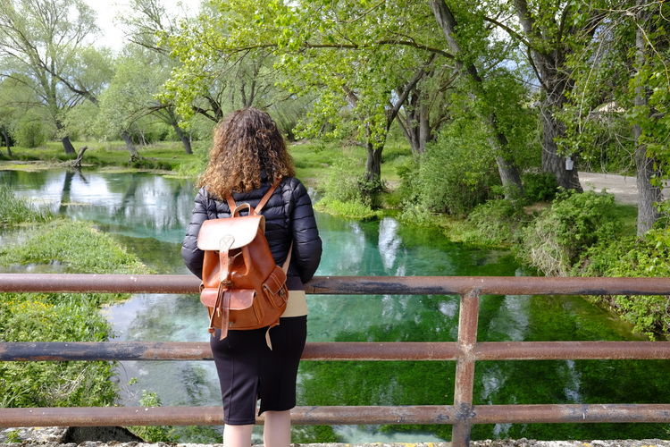 Rear view of woman with backpack standing on footbridge over river against trees