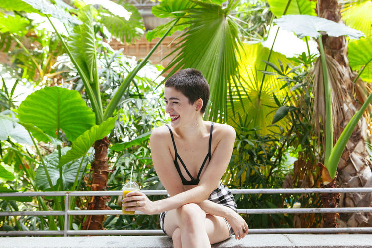 Smiling young woman with short hair holding juice while sitting against plants