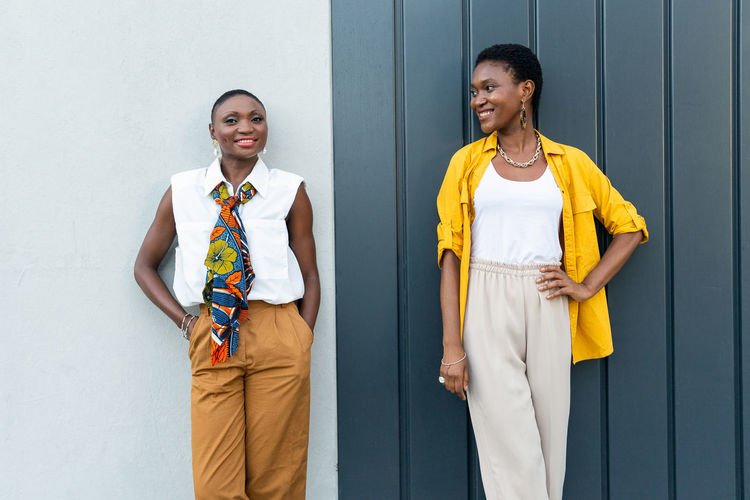 Confident adult african american female friends with short dark hair in stylish outfits smiling and looking at camera while standing against gray wall in city