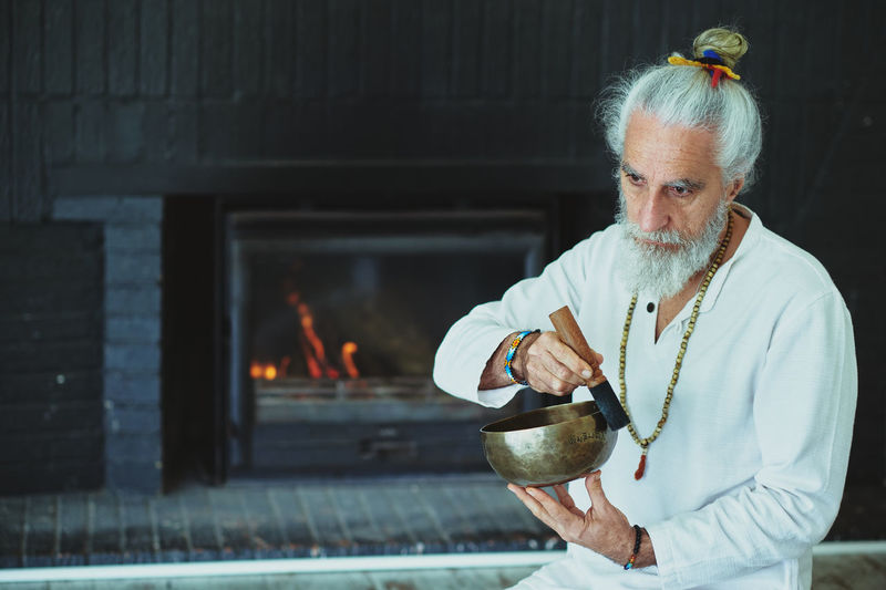 Elderly male with gray beard playing singing bowl with wooden striker while looking away during spiritual practice